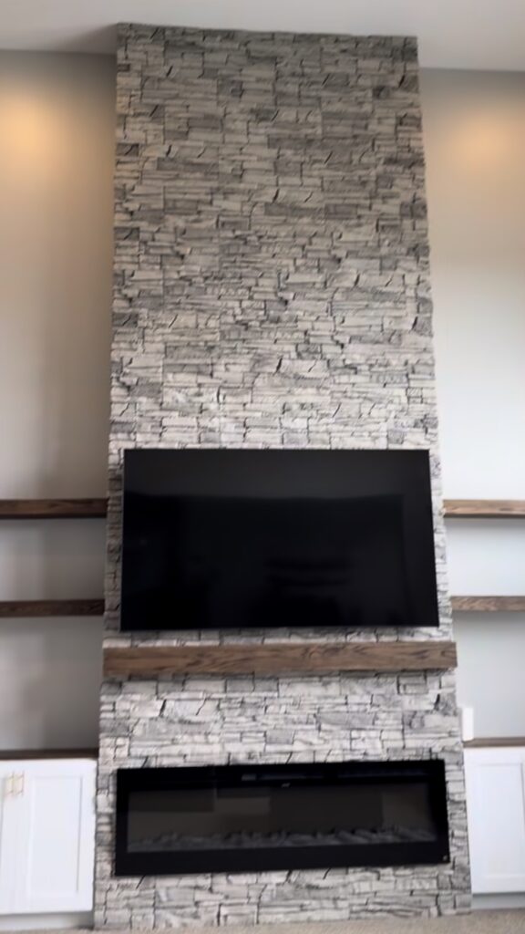 An 8-foot fireplace wall built with faux stacked stone, custom white shaker cabinets, oak floating shelves, and a Touchstone electric fireplace. See how Radford Woodworks built a full-height focal point that transformed this Indiana living room.