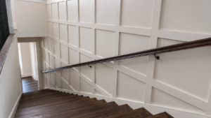 Square board and batten grid wall running down a two-story stairwell with a wall-mounted dark handrail and dark hardwood treads