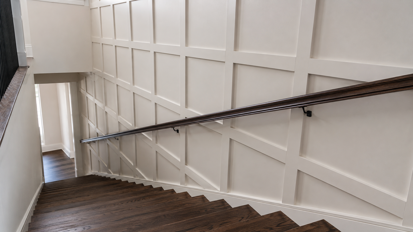 Square board and batten grid wall running down a two-story stairwell with a wall-mounted dark handrail and dark hardwood treads