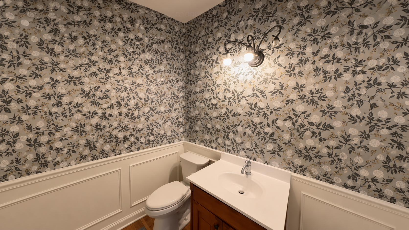 Charcoal botanical wallpaper with white blooms over white wainscoting in a central Indiana powder room with a wood vanity and bronze sconce by Radford Woodworks